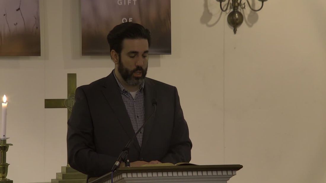 Pastor Adam Miller preaching from the pulpit in the sanctuary, with a brass cross and candle beside him